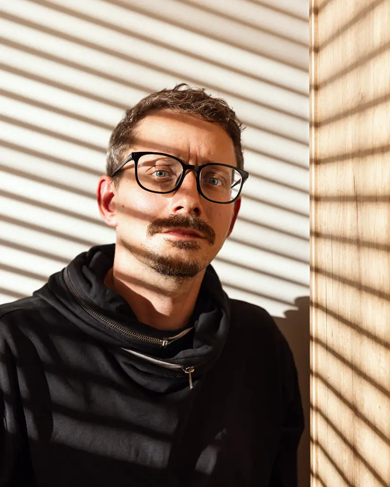 Portrét muže s brýlemi a knírkem. Světlo a stín z žaluzií. Černá mikina.