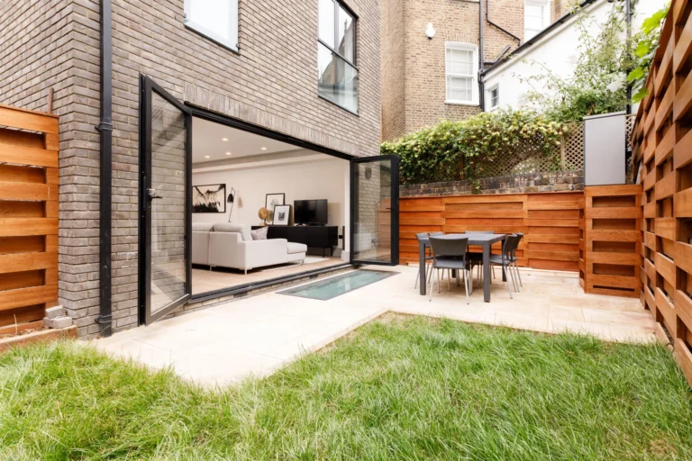 Moderní dům s terasou a zahradou. Zelený trávník, dřevěný plot, jídelní stůl a otevřené dveře do obýváku. Architektura.